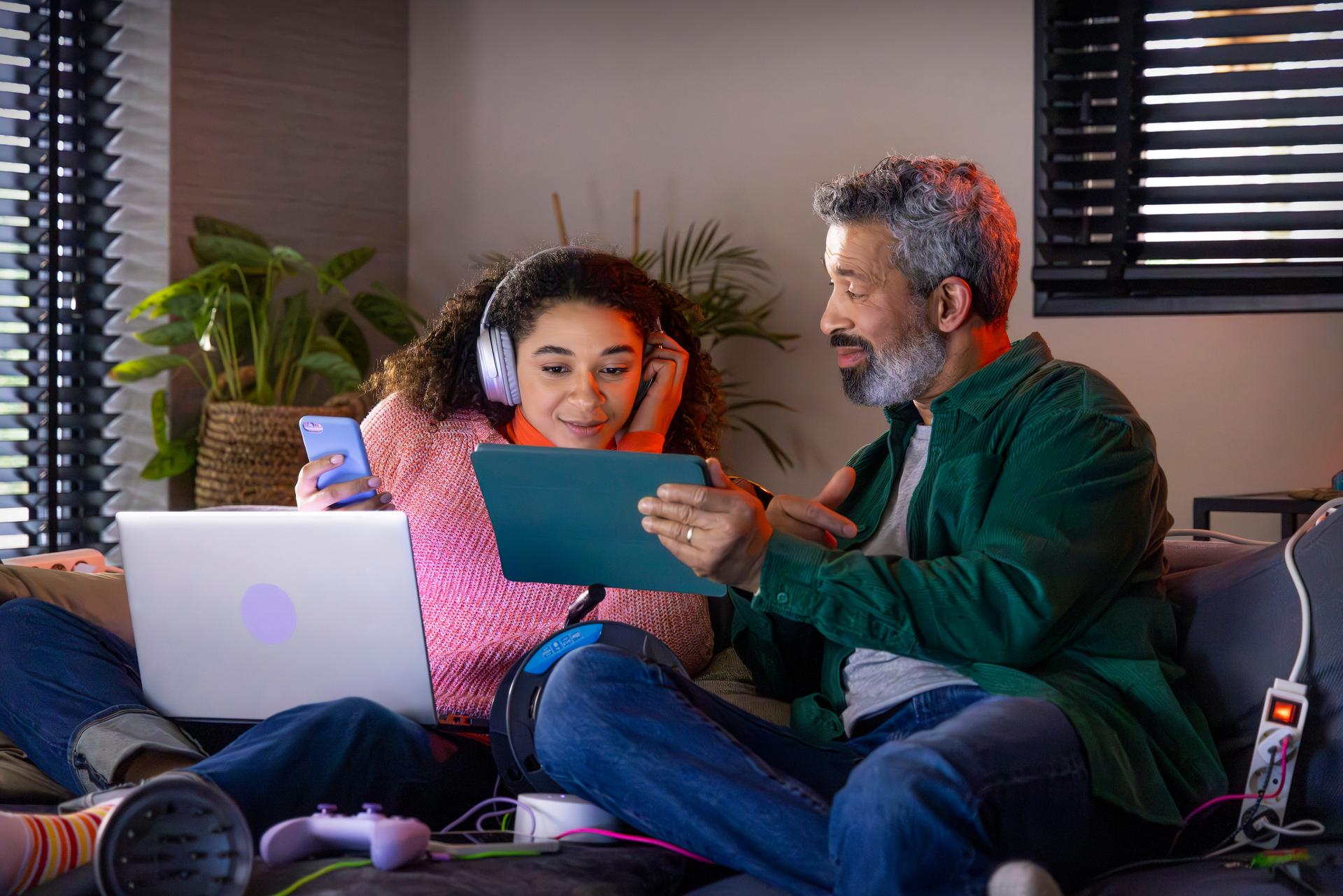 Vader en dochter zitten op de bank. Vader laat aan dochter iets zien op de tablet.