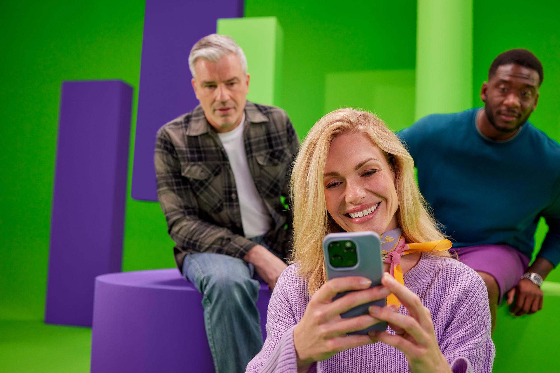 Vrouw kijkt op haar telefoon. Twee mannen die achter haar zitten kijken mee. 