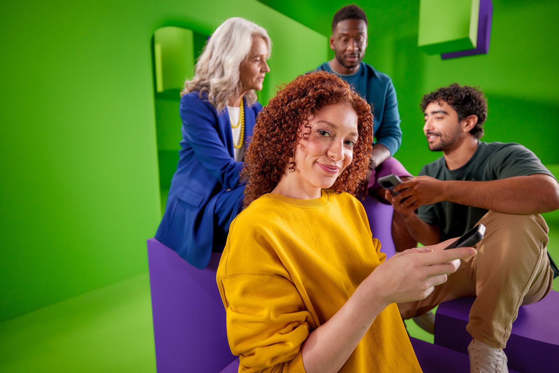 Vrouw houdt telefoon in haar handen en kijkt de camera in. Achter haar kijken 3 mensen samen op een telefoon.
